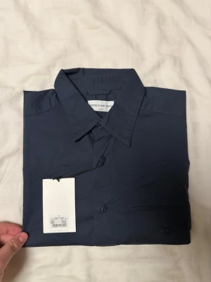 Mörkblå skjorta från These Glory Days - Stilren mörkblå skjorta från These Glory Days med klassisk krage och knappstängning framtill. Skjortan har en bröstficka med lock och är tillverkad i bomull, perfekt för en clean och enkel look.