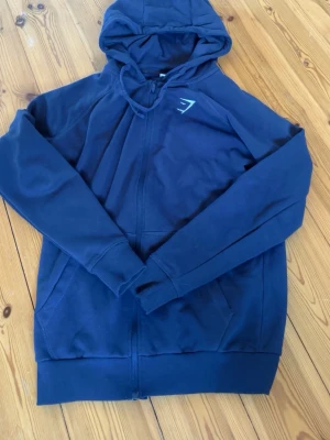 Marinblå hoodie från Gymshark - Snygg marinblå hoodie från Gymshark med dragkedja framtill och två fickor. Hoodien har en broderad vit Gymshark-logga på bröstet och är tillverkad i mjukt bomullsmaterial. Perfekt för chill eller träning.