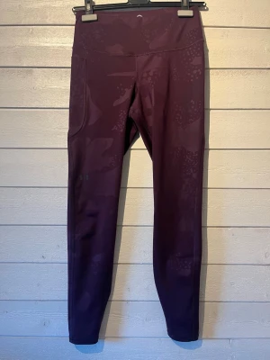 Mörklila leggings med mönster fr Under Armour  - Säljer ett par plommonfärgade tights med diskret mönster. De har hög midja och är tillverkade i ett stretchigt, mjukt material som sitter skönt på kroppen. Perfekta för träning eller chill. Inga synliga loggor eller fickor. Strl M . 
