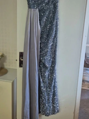 Glittrig silver långklänning - Säljer en snygg långklänning i silver med glittriga paljetter och en axelbandslös design. Klänningen har en elegant slits och en del i slätt tyg som kontrast till paljetterna. Perfekt för dig som vill sticka ut på bal eller fest!