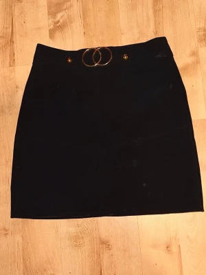 Svart kortkjol med ringdetalj - Stilren svart kortkjol med dekorativ ringdetalj i midjan och dragkedja bak. Kjolen har en enkel, rak passform och är perfekt att matcha med olika toppar. Materialet känns mjukt och har en lätt struktur.