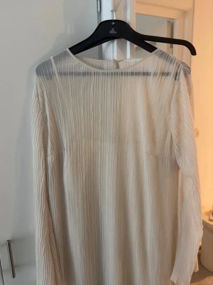 Vit långärmad plisserad klänning - Säljer en vit långärmad klänning med plisserad struktur och rund hals. Klänningen har en rak passform och är tillverkad i ett lätt, transparent material som ger ett luftigt intryck. Perfekt för dig som gillar minimalistisk och stilren design. Använd en gång 