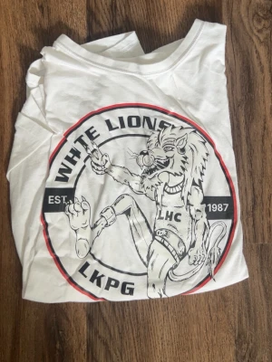Vit White Lions t-shirt LHC - Säljer en vit t-shirt från White Lions med stort tryck av en tecknad lejonmaskot och texten LHC LKPG EST. 1987 på framsidan. Klassisk rund hals och korta ärmar. 
