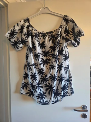 Vit sommarklänning med palmmönster - Säljer en luftig vit klänning med svart palmmönster, perfekt för sommaren. Klänningen har korta ärmar, offshoulder-modell och avslappnad passform. Tillverkad i bomull för en skön känsla. Superfin till stranden eller cityhänget!