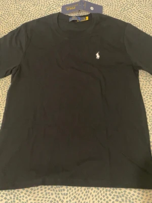 Svart t-shirt från Polo Ralph Lauren - Klassisk svart t-shirt från Polo Ralph Lauren med rund hals och korta ärmar. T-shirten har den ikoniska vita Polo-loggan broderad på bröstet och är tillverkad i mjuk bomull för en skön känsla. Perfekt till jeans eller shorts.