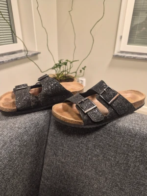 Svarta glittriga sandaler med spännen från Colors Of California 41 - Säljer ett par svarta sandaler med två breda remmar i glittrigt material och justerbara metallspännen. Fotbädden är i kork-look och sulan är platt. Perfekta för dig som vill ha både komfort och stil.