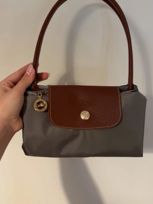 Grå och brun handväska från Longchamp - Snygg handväska från Longchamp i grå nylon med bruna detaljer i läder. Väskan har ett guldfärgat dragkedjesmycke med Longchamp-loggan och ett lock med tryckknapp. Perfekt storlek för det viktigaste och enkel att matcha till olika outfits.