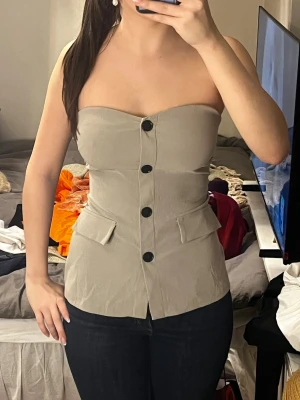 Beige/grön axelbandslös korsettopp - Trendig beige/grön korsettopp med stora svarta knappar framtill och fickdetaljer vid midjan. Axelbandslös design som ger en clean och stilren look. Sitter tight och formar kroppen snyggt. Perfekt att styla med jeans eller kjol.