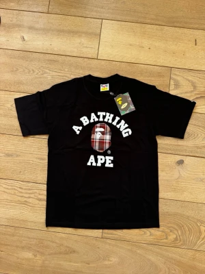 Svart A Bathing Ape t-shirt - Säljer en svart t-shirt från A Bathing Ape med det klassiska BAPE-huvudet i röd och vit rutig design på bröstet. T-shirten har vit text och en normal passform. Perfekt för dig som gillar streetwear och vill sticka ut.