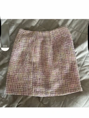 Rutig kortkjol i pastellfärger - Trendig kortkjol med rutigt mönster i rosa, lila, vitt och lite grönt. Kjolen har en rak passform och en liten slits bak för extra rörelsefrihet. Materialet känns som en blandning av bomull och polyester, vilket ger en lätt struktur.