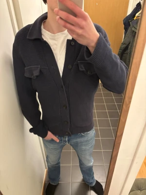 Mörkblå kofta från Dobber - Snygg mörkblå kofta från Dobber med knäppning framtill och två bröstfickor med lock. Grovstickad struktur och klassisk krage ger en avslappnad look. Perfekt att slänga över en t-shirt för en chill stil.