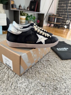Svarta Golden Goose sneakers med silverdetaljer - Golden Goose sneakers i svart mocka med ikonisk silverfärgad stjärna på sidan och matchande silverdetalj vid hälen. Skorna har beige snörning och en klassisk låg profil. Snygg kontrast mellan material och färger, perfekt för dig som gillar streetstyle.