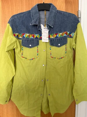  skjorta  - Skjorta  limegrön tyg med blå jeansdetaljer upptill och på fickorna. brodyr längs bröstet och fickkanterna. Perfekt för dig som gillar färg och mönster. Har inte använts, helt ny 