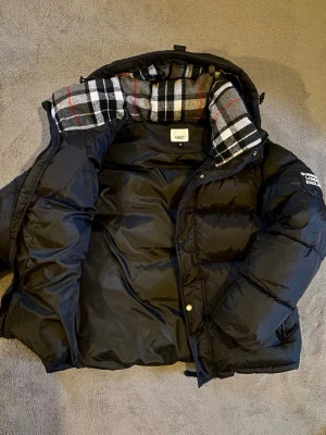 Svart dunjacka från Beyond Retro - Snygg svart dunjacka från Beyond Retro med rutigt foder i huvan. Jackan har en puffig passform, dragkedja och knappar framtill samt en huva som ger extra värme. Perfekt för kalla vinterdagar och riktigt skön att bära.