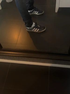 Mörkblå Adidas sneakers med vita ränder - Snygga mörkblå sneakers från Adidas med klassiska vita ränder på sidorna och vit sula. Skorna har en låg profil och är tillverkade i mockaliknande material med textildetaljer. Perfekta för en avslappnad och sportig stil. Storlek 43,44