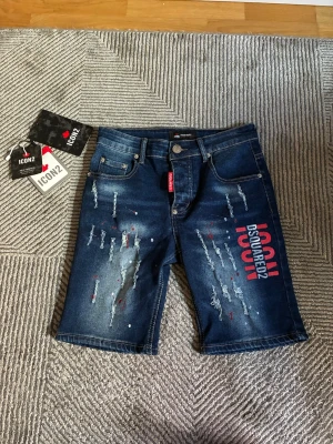 Blå jeansshorts Dsquared2 ICON - Säljer ett par blå jeansshorts från Dsquared2 ICON med slitningar och färgstänk. Shortsen har tryckt logga i vitt och rött på ena benet, klassisk femficksmodell och knappgylf. Materialet är denim i bomull och de har en cool streetwear-vibe.