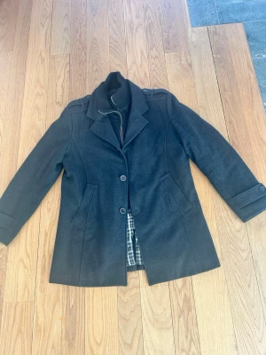 Svart rock från Bark of Sweden - Stilren svart rock från Bark of Sweden med klassisk krage, två knappar framtill och axelklaffar. Fodrad med rutigt innerfoder och har två snedställda fickor. Perfekt för kyligare dagar och ger en snygg siluett till din outfit. Ena innerfickan är trasig men kan enkelt åtgärdas och är inte synligt utifrån.