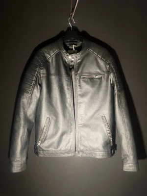 Svart Skinnjacka - Snygg svart skinnjacka med raka linjer och modern passform. Jackan har ståkrage med tryckknapp, tre dragkedjeförsedda fickor och diskreta sömdetaljer på axlarna. Perfekt för dig som gillar en stilren och tidlös look.