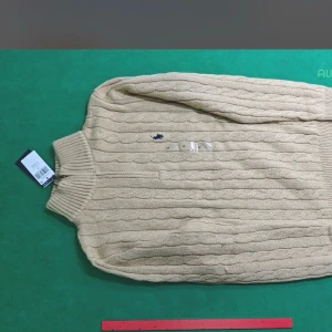 Beige kabelstickad tröja Polo Ralph Lauren - Kabelstickad beige tröja från Polo Ralph Lauren med hög krage och broderad marinblå logga på bröstet. Tröjan har långa ärmar och ribbade muddar vid hals, ärmslut och nederkant. Perfekt för lager på lager och klassisk stil.