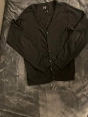 Mörkgrå cardigan från Faded - Stilren mörkgrå cardigan från Faded med v-ringning och knäppning framtill. Tillverkad i mjukt material och har långa ärmar samt ribbade muddar vid ärmslut och nederkant. Perfekt att slänga över en t-shirt för en avslappnad look.