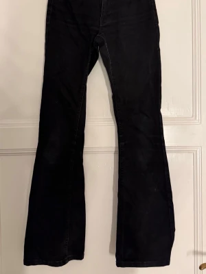 Svarta bootcut jeans från Ginatricot - Säljer ett par svarta bootcut jeans från Ginatricot. De har låg-medel hög midja, utsvängda ben och klassiska fem fickor. Jeansen är i ett stretchigt denimtyg som sitter snyggt och bekvämt. Perfekta för dig som gillar en retroinspirerad look. Lite urtvättade men inge mer synligt slitage.
