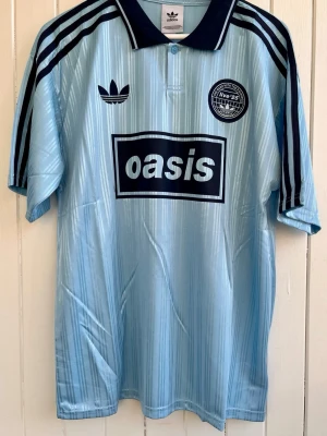 Oasis - Turné 25 - Fotbollströja  - Säljer en ljusblå  Oasis-supportertröja för bandets turné under 2025. Tröjan är en reprint i nyskick. Aldrig använd. ”25” på ryggen.      Storlek: Large 