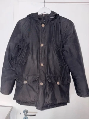 Woolrich Down Parka/ Dunjacka - Snygg svart parkas från Woolrich med huva och stora fickor framtill. Jackan har dragkedja och knappar, samt en varm beige innerfoder. Sparsamt använd Såklart äkta passar som standard M Pris kan diskuteras vid snabb affär.