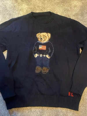 Mörkblå stickad tröja med björnmotiv - Säljer en mörkblå stickad tröja från Ralph Lauren med det ikoniska björnmotivet på bröstet. Björnen har en tröja med amerikanska flaggan och detaljerade broderier. RL-broderi i rött vid nederkanten. Klassisk rund hals och ribbade muddar. (Rep)