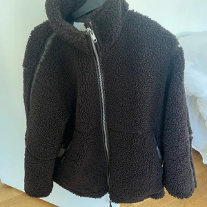 Brun teddyjacka från HM XS - Mysig mörkbrun teddyjacka med hög krage och hel dragkedja framtill. Jackan har två stora fickor och är gjord i fluffigt teddy-material som ger en riktigt skön och trendig look. Den har en liten flint-klump men syns ej då armen är ivägen. Kan lätt fixas
