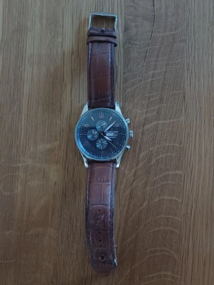 Jacques Lemans kronograf klocka brun - Stilren kronograf från Jacques Lemans med mörkblå urtavla, silverfärgat boett och brunt läderarmband med krokopräglat mönster. Klockan har flera subdials och tydliga markeringar. Perfekt accessoar för dig som gillar klassisk design. Bandet går att ändra men klockan är som ny.