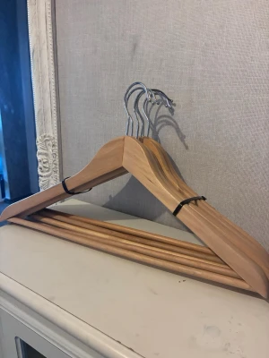 Trägalgar med metallkrok 6st - Säljer ett set med 6 stilrena galgar i ljust trä med metallkrok. Perfekta för att hänga upp kläder i garderoben och håller formen på skjortor, jackor eller tröjor. Galgarna har en klassisk design. Bra skick!!