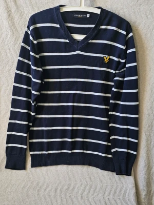 Randig stickad tröja Lyle & Scott - Snygg svart stickad tröja från Lyle & Scott med vita horisontella ränder och gul logga på bröstet. V-ringad modell med ribbade muddar vid ärmslut och nederkant. Perfekt för dig som gillar klassisk och stilren design.