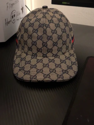 GG keps med GG-mönster - Säljer en beige keps från GG med klassiskt GG-monogram i mörkblått över hela kepsen. Kepsen har böjd skärm och röda samt gröna ränder på sidorna. Tillverkad i canvasmaterial och har en modern streetwear-look.