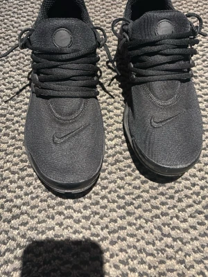 Svarta Nike prestos - Säljer ett par helsvarta Nike sneakers med lågt skaft. Skorna har en stilren design med diskret Nike-logga på sidan och ovandel i textilmaterial. Klassiska svarta skosnören och bekväm passform. Perfekta för dig som gillar en clean look.