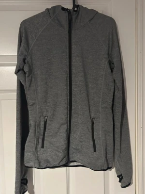 Träningströja - Grå sportjacka med dragkedja framtill och två fickor med svarta dragkedjor. Jackan har huva, långa ärmar och praktiska tumhål som ger extra komfort. Perfekt för träning, löpning eller en aktiv vardag.(har nån liten tråd lös)ifrån h&m) 