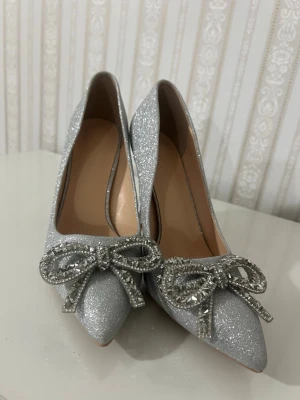Silvriga glittriga pumps med rosett - Snygga silvriga pumps med glitter och spetsig tå. Skorna har en dekorativ rosett framtill täckt av glittrande stenar som ger extra bling. Klackarna är smala och eleganta, perfekta för att sticka ut. Materialet är glittrigt tyg och insidan är beige.