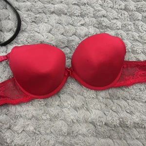 Röd push-up bh med spetsdetaljer - Snygg röd push-up bh med vadderade kupor och spetsdetaljer längs sidorna. Bh:n har justerbara axelband och hakknäppning bak. Materialet är en mix av slät textil och spets som ger en feminin look.