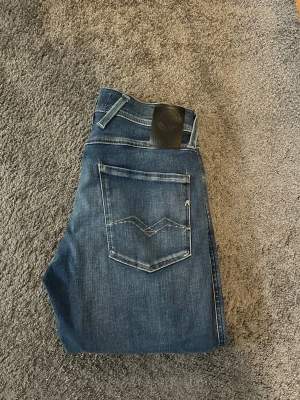 Blå jeans från Replay  - Säljer ett par klassiska blå jeans från Nudie Jeans med raka ben och snygga slitningar framtill. Jeansen har fem fickor, kontrastsömmar och en svart patch med logga bak i midjan. Perfekta för en avslappnad och trendig look.