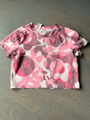 Rosa mönstrad t-shirt från Adidas - Säljer en kortärmad t-shirt från Adidas med ett unikt abstrakt mönster i olika rosa och lila toner. T-shirten har en croppad passform och är tillverkad i mjuk bomull, perfekt för en trendig och avslappnad stil.