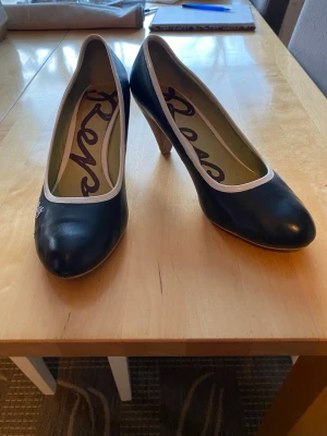 Svarta pumps från Replay - Snygga svarta pumps från Replay med vit kant runt öppningen och broderad logga på sidan. Skorna har rundad tå och klack i trälook. Tillverkade i slätt läderliknande material. Perfekta för dig som vill ha en klassisk men ändå unik stil.