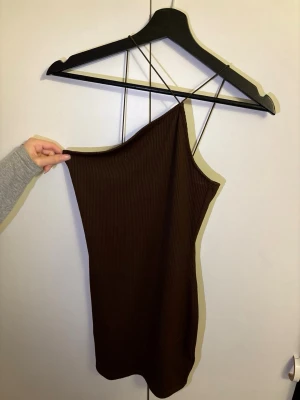 Brun ribbad one shoulder klänning - Säljer en brun, ribbad kortklänning med one shoulder-design och tunna axelband. Klänningen har en tight passform. Materialet känns mjukt och stretchigt, vilket gör den bekväm att bära.