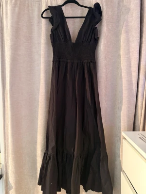 Svart långklänning från Boohoo - Snygg svart långklänning från Boohoo med djup v-ringning och breda axelband. Klänningen har smock i midjan för en bekväm passform och volangdetalj längst ner. Klänningen är oanvänd, prislappen sitter kvar.