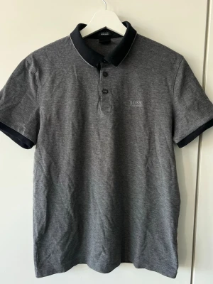 Grå pikétröja från BOSS - Snygg grå pikétröja från BOSS med svart krage och knappslå. Tillverkad i bomull. Nypris: 1189kr 