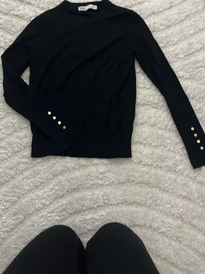 Svart tröja från Zara med gulddetaljer - Snygg svart långärmad tröja från Zara med rund halsringning och fyra guldfärgade knappar på ärmsluten. Tröjan har en smal passform och är tillverkad i ett mjukt, stretchigt material som sitter skönt på kroppen.