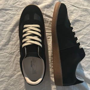 Svarta maison gats  - Snygga svarta sneakers med vita snören och klassisk gummisula i brunt. Skorna har en stilren design med svarta paneler och en låg siluett. Insidan är ljus och materialet ser ut att vara en mix av mocka och läder. Perfekta för en clean och modern look.