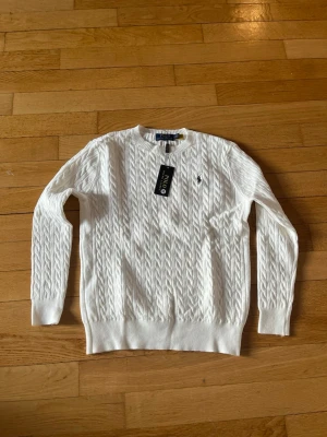 Vit kabelstickad tröja Polo Ralph Lauren - Vit kabelstickad tröja från Polo Ralph Lauren med rund halsringning och den klassiska broderade loggan på bröstet. Tröjan har långa ärmar och ribbade muddar vid ärmslut och nederkant. Perfekt för dig som gillar stilrena och tidlösa plagg.