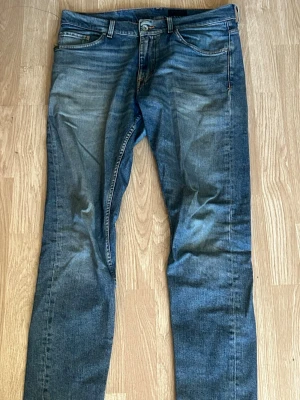 Blå raka jeans från Tiger Of Sweden - Säljer ett par blå jeans med raka ben och snygga slitningar framtill. Jeansen har klassisk femficksdesign, knappgylf och kontrastsömmar. Materialet är denim i bomull och passformen är relaxed fit. Perfekta för en avslappnad och trendig look.