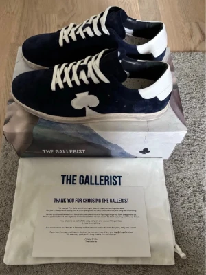 Mörkblå sneakers från The Gallerist - Snygga mörkblå sneakers i mocka från The Gallerist med vita detaljer, vita skosnören och en unik vit klöver på sidan. Skorna har vit sula och hälparti, vilket ger en stilren kontrast. Perfekta för dig som vill sticka ut med en cool och modern look.