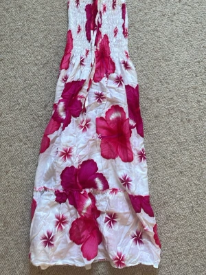Vit och rosa blommig långklänning - Säljer en somrig långklänning med stora rosa blommor på vit botten. Klänningen har smock upptill och tunna axelband som knyts i nacken. Perfekt för varma dagar och ger en avslappnad, bohemisk vibe.