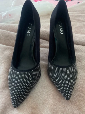 Svarta pumps med strass från Tiamo - Snygga svarta pumps från Tiamo med spetsig tå och täckt häl. Skorna är dekorerade med glittrande strass framtill som ger en lyxig känsla. Materialet ser ut att vara textil med en mjuk finish. Perfekta för dig som vill sticka ut lite extra.